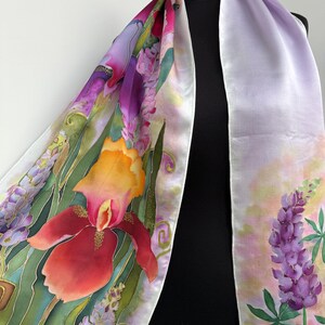 K&ouml;nnte beinhalten: Ein langer Seidenschal mit handgemaltem Blumenmuster, das leuchtend rote und orangefarbene Schwertlilien, violette Lupinen und gr&uuml;nes Blattwerk vor einem zarten lavendelfarbenen und wei&szlig;en Hintergrund zeigt. Der Stoff hat eine glatte, gl&auml;nzende Oberfl&auml;che.
