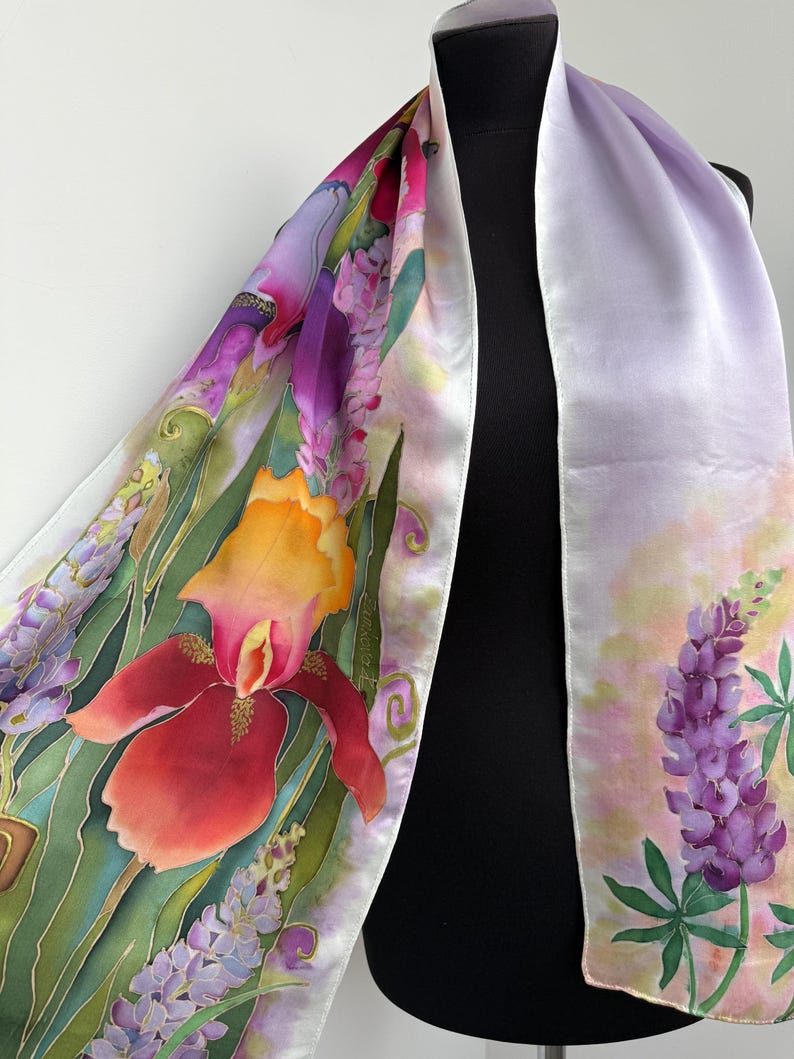 K&ouml;nnte beinhalten: Ein langer Seidenschal mit handgemaltem Blumenmuster, das leuchtend rote und orangefarbene Schwertlilien, violette Lupinen und gr&uuml;nes Blattwerk vor einem zarten lavendelfarbenen und wei&szlig;en Hintergrund zeigt. Der Stoff hat eine glatte, gl&auml;nzende Oberfl&auml;che.