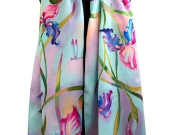 May include: A silk scarf featuring a watercolor design of iris flowers in shades of pink, purple, and blue. The scarf has a light blue and lavender background with green stems and a dragonfly. The scarf is draped over a black form.