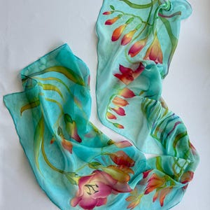 Pode incluir: Um len&ccedil;o leve e semitransparente num tom azul turquesa vibrante, com um design floral pintado &agrave; m&atilde;o. O padr&atilde;o apresenta folhas verdes delicadas e alongadas, al&eacute;m de cachos de flores desabrochadas em tons de rosa, laranja e amarelo.