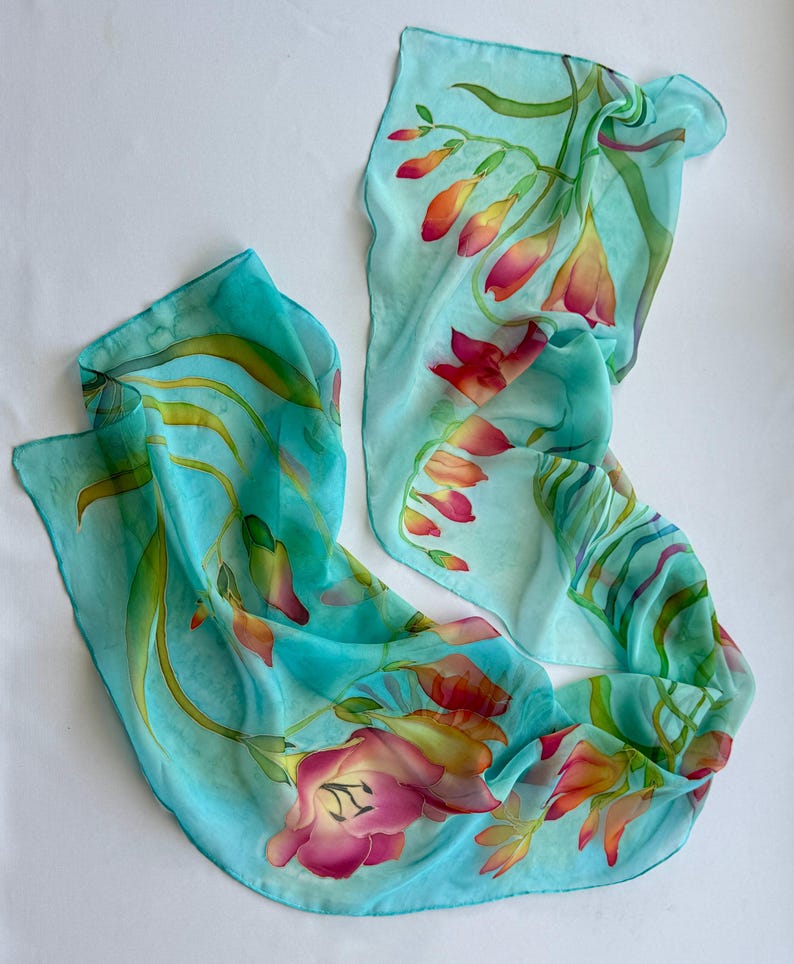 Pode incluir: Um len&ccedil;o leve e semitransparente num tom azul turquesa vibrante, com um design floral pintado &agrave; m&atilde;o. O padr&atilde;o apresenta folhas verdes delicadas e alongadas, al&eacute;m de cachos de flores desabrochadas em tons de rosa, laranja e amarelo.