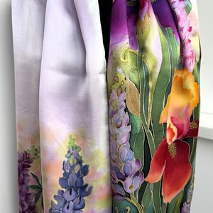 K&ouml;nnte beinhalten: Ein langer rechteckiger Schal mit handgemaltem Blumenmuster. Eine Seite zeigt einen zarten lavendelfarbenen Hintergrund mit blauen Lupinen. Die andere Seite pr&auml;sentiert leuchtend gr&uuml;nes Blattwerk, violette Schwertlilien, orange Lilien und eine kleine braune Schnecke.