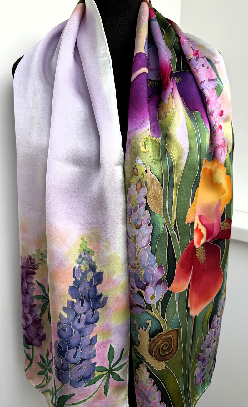 K&ouml;nnte beinhalten: Ein langer rechteckiger Schal mit handgemaltem Blumenmuster. Eine Seite zeigt einen zarten lavendelfarbenen Hintergrund mit blauen Lupinen. Die andere Seite pr&auml;sentiert leuchtend gr&uuml;nes Blattwerk, violette Schwertlilien, orange Lilien und eine kleine braune Schnecke.