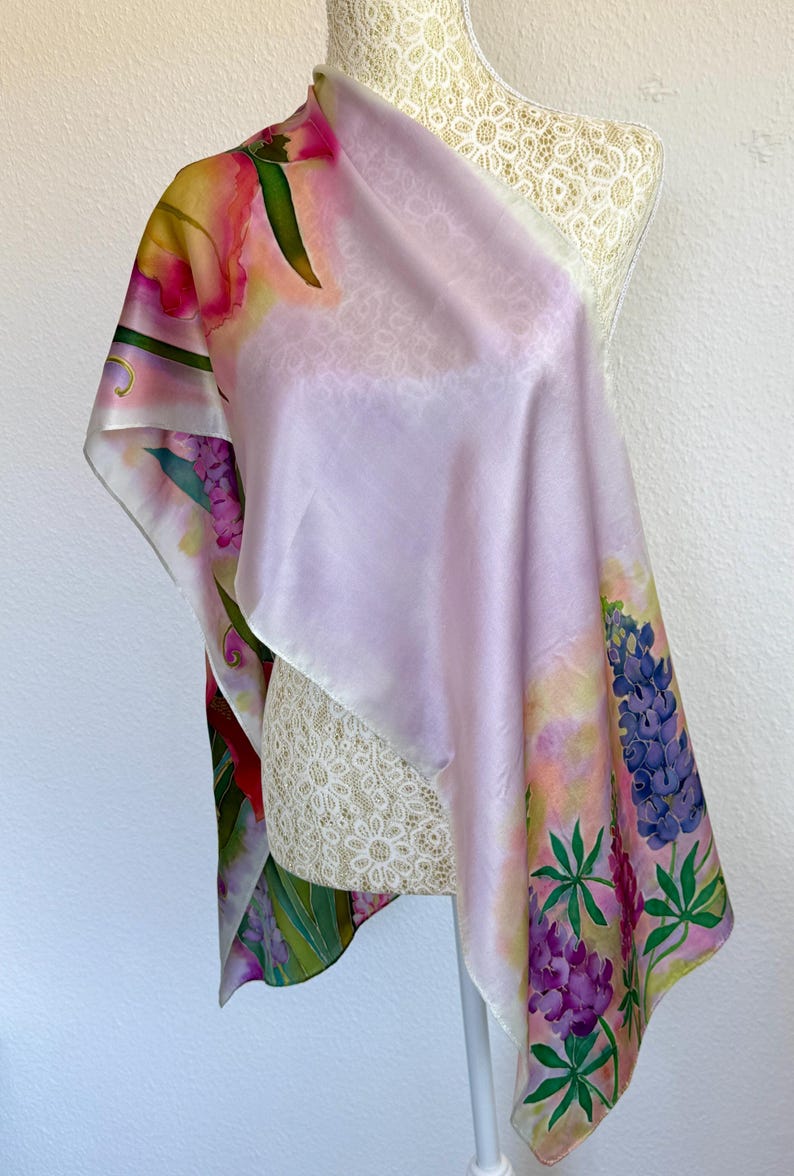 K&ouml;nnte beinhalten: Ein quadratischer Seidenschal mit einem sanften lavendelfarbenen Zentrum und einem lebendigen Blumenrand. Das Design zeigt handgemalte Illustrationen von violetten Lupinen, gr&uuml;nen Bl&auml;ttern und rosa Bl&uuml;ten auf einem hellen, pastellfarbenen Hintergrund.
