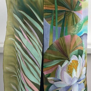 Puede incluir: Una bufanda de seda verde y azul con un dise&ntilde;o pintado a mano de nen&uacute;fares y hojas. La bufanda est&aacute; doblada y drapeada sobre el cuello de un maniqu&iacute;.