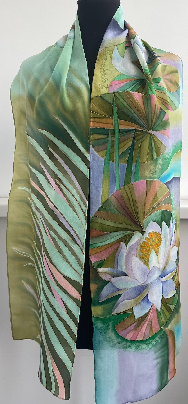 Puede incluir: Una bufanda de seda verde y azul con un dise&ntilde;o pintado a mano de nen&uacute;fares y hojas. La bufanda est&aacute; doblada y drapeada sobre el cuello de un maniqu&iacute;.