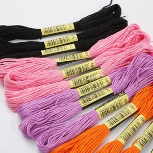 May include: A collection of embroidery floss skeins in black, pink, purple, and orange. Each skein is secured with a paper band featuring a barcode and the color numbers 310, 604, 209, and 740, arranged in a fan-like pattern on a white surface.