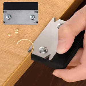 May include: A handheld woodworking edge trimmer featuring a black plastic body and a sharp metal blade secured by two silver screws. The tool is shown shaving a thin layer of wood from the edge of a light brown wooden board, creating small curls.