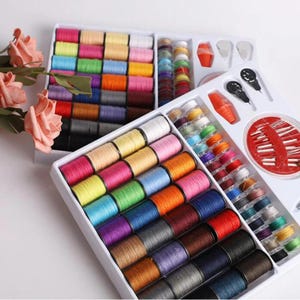 Può includere: Un kit da cucito con una varietà di rocchetti di filo colorati, aghi e altri accessori per cucire. Il kit include rocchetti di filo in vari colori, tra cui rosa, giallo, blu e marrone. Il kit è ben organizzato in un contenitore di plastica bianco.