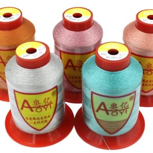 May include: Five spools of AOYI brand embroidery thread in orange, pink, copper, silver, and teal. Each spool features a red plastic base and top, with a yellow label displaying the AOYI logo and Chinese characters. The thread is wound tightly.