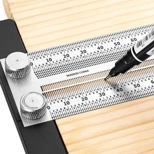 May include: A metal woodworking marking gauge with a black base and a silver ruler featuring metric measurements up to 60 cm. The tool includes two silver knurled knobs and a black marker pen tip positioned within the central slotted guide.