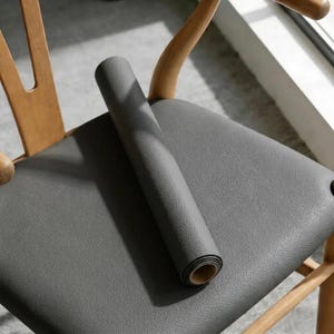 May include: A rolled-up grey mat with a textured surface resting on a matching grey seat cushion. The mat features a consistent pebbled grain pattern across its exterior, while the inner core of the roll reveals a smooth, light-coloured circular edge.