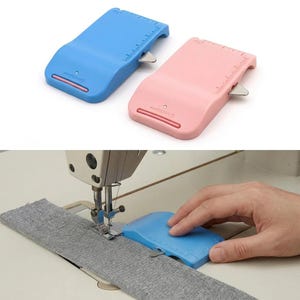 May include: A light blue and a light pink sewing seam guide tool, each featuring a built-in metal blade and integrated ruler markings. The blue tool is shown in use on a sewing machine, positioned against a strip of grey fabric to guide stitching.