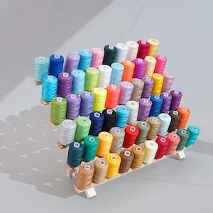 Könnte beinhalten: Ein abgestuftes Holzregal mit einer großen Sammlung von Nähgarnspulen in einer Vielzahl von Farben, darunter Blau, Grün, Pink, Gelb, Orange, Lila, Rot, Weiß, Schwarz, Braun und Grau, die in ordentlichen Reihen auf einem hellen Holzständer stehen.