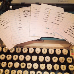 May include: A vintage typewriter with a stack of index cards with handwritten quotes on them. The quotes are about love, life, and summer. The typewriter is black and has a white keyboard.