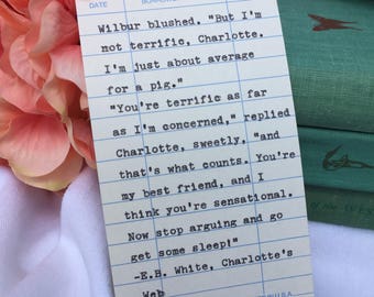 Puede incluir: Una tarjeta de biblioteca rectangular con un extracto impreso de Charlotte's Web de E.B. White. La tarjeta tiene líneas horizontales azules y encabezados para la fecha y el nombre del prestatario, con el texto en fuente de máquina de escribir.