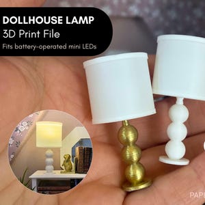 May include: Two miniature dollhouse lamps with white shades. One lamp has a gold base, the other a white base. The lamps are held in a hand. Text on the image reads "DOLLHOUSE LAMP 3D Print File Fits battery-operated mini LEDs".