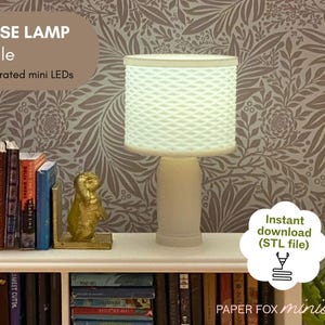 May include: A miniature dollhouse lamp with a textured white lampshade and base. The lamp is next to a shelf with miniature books and a gold-colored bookend. Text on the image reads "DOLLHOUSE LAMP 3D Print File Fits battery-operated mini LEDs".