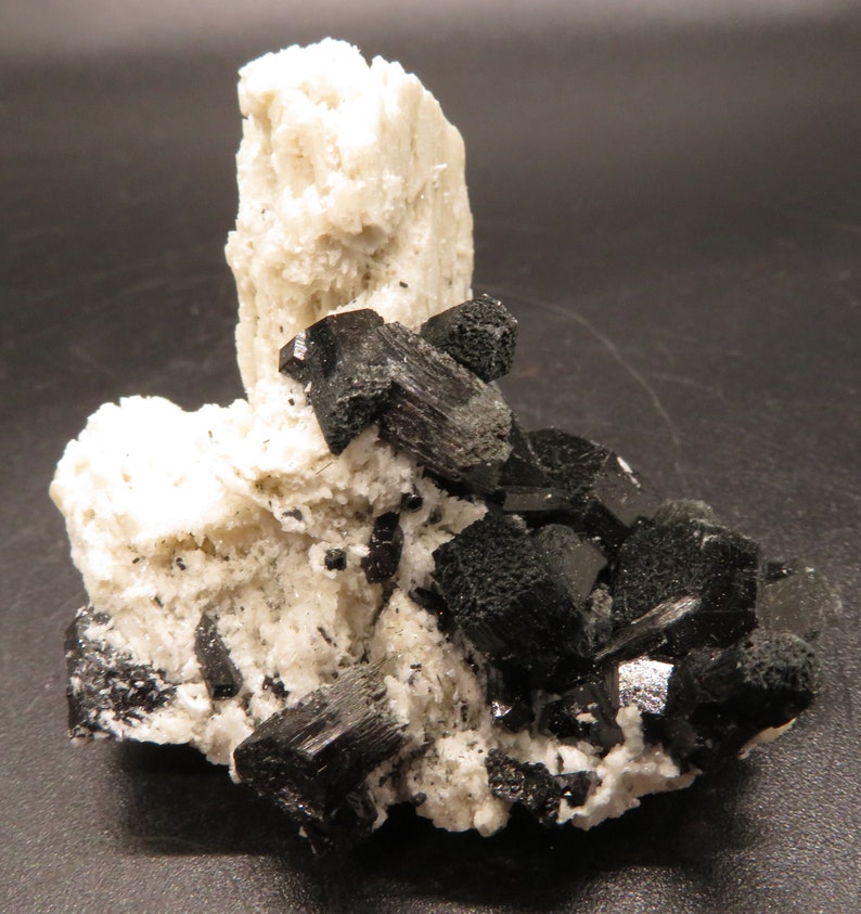 May include: A cluster of black crystals on a white mineral matrix. The crystals are sharp and well-defined, with a glossy finish. The matrix is rough and textured.