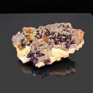 May include: A close-up of a raw mineral specimen. The stone features a mix of colors, including purple, orange, green, and beige. The mineral has a rough, textured surface and is displayed against a dark background.