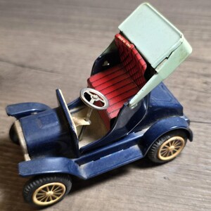 May include: A vintage-style blue tin toy car featuring a red textured seat, a pale green convertible top, and a silver steering wheel. The wheels are yellow with black tires, and the text "MADE IN JAPAN" is printed on the red seat back.