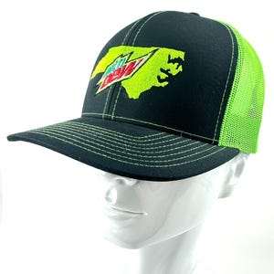 Puede incluir: Gorra de camionero negra con la parte trasera y la visera de malla verde neón. La parte delantera presenta un diseño bordado de un contorno verde de un estado con el logotipo de Mountain Dew. La gorra tiene costuras verde neón.