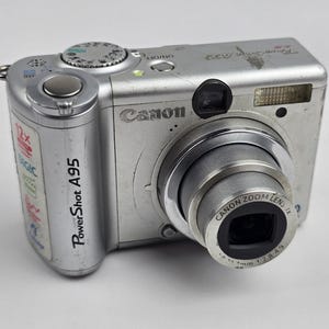 May include: A silver Canon PowerShot A95 digital camera featuring a prominent lens barrel, a shutter button, a mode dial on top, and a grip area on the left side. The front body displays the Canon logo, a flash unit, and various technical labels.