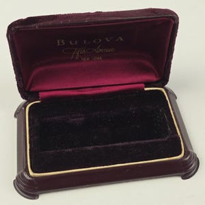Może przedstawiać: Otwarta, prostokątna szkatułka na biżuterię Bulova Fifth Avenue. Pudełko jest w kolorze ciemnego bordo z pluszowym, aksamitnym wnętrzem i złotą obwódką. Logo Bulova jest widoczne wewnątrz wieczka.