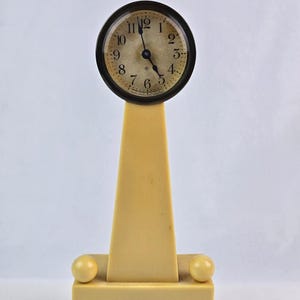 Puede incluir: Un reloj antiguo con esfera redonda y marco negro, colocado sobre una base geométrica de color crema. La esfera del reloj muestra números y manecillas negras. La base tiene una parte inferior rectangular con dos acentos esféricos.