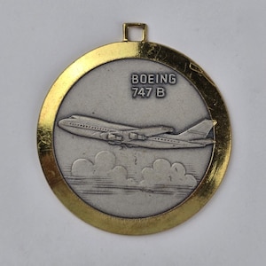 May include: A round, silver-colored pendant with a gold-colored rim and a small loop at the top. The pendant features an image of a Boeing 747 B airplane flying above clouds, with the text "BOEING 747 B" above the plane.