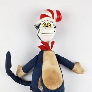 May include: A vintage plush toy of the Cat in the Hat from the children's book by Dr. Seuss. The toy is wearing a red and white striped hat, a red bow tie, and a blue and brown outfit. The toy has a long tail and is in good condition, but shows signs of wear.