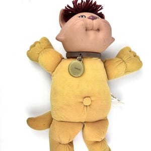 Peut inclure: Poupée chat Cuddleez vintage avec un visage beige et un corps jaune. Le chat en peluche porte un collier marron avec une étiquette dorée « Cuddleez ». Il a des cheveux en fil marron et est dans une pose ludique, les bras levés.