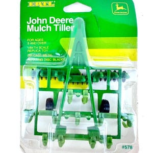 May include: A green John Deere Mulch Tiller toy in its original packaging. The package includes the text "John Deere Mulch Tiller", "1/64th scale replica toy", and "rotating disc blades". The toy is made of die-cast metal and is for ages 3 and over.