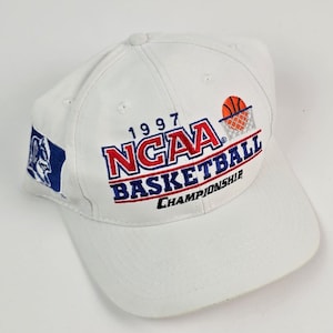 Może przedstawiać: Biała czapka bejsbolowa z napisem "1997 NCAA BASKETBALL CHAMPIONSHIP" w kolorze czerwonym i niebieskim. Czapka ma grafikę koszykówki i siatki oraz niebiesko-białe logo z boku.