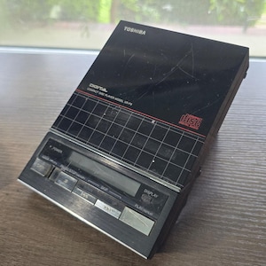 May include: A black Toshiba Digital Compact Disc Player, model XR-P9, with a grid pattern on the top. The player has a silver control panel with buttons for power, play/pause, skip, and display. The "Digital Audio" logo is visible.