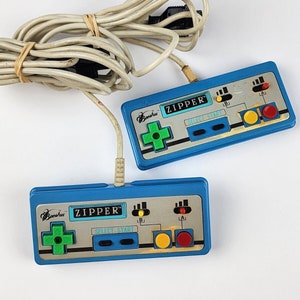 Könnte beinhalten: Zwei blaue und graue Videospiel-Controller mit dem Text "ZIPPER" darauf. Die Controller haben ein grünes Steuerkreuz, zwei schwarze Tasten mit der Aufschrift "SELECT" und "START" sowie drei Tasten mit der Aufschrift "A", "B" und "LA".