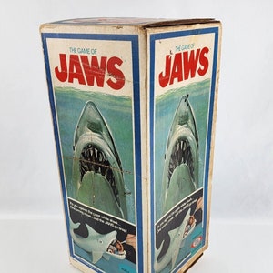 May include: A vintage board game box for the game "Jaws" featuring a large image of the great white shark with its mouth open. The box is blue and white with the game title in red letters. The text on the box reads "It's you against the great white shark... One wrong move... and the JAWS go snap!"
