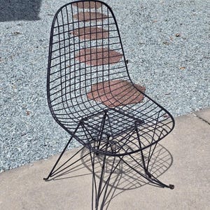 May include: Black wireframe chair with a rounded back and a seat made of a grid pattern. The chair has four thin, angled legs. The chair is sitting on a concrete surface.