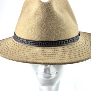 May include: A tan fedora hat with a dark brown leather band. The hat has a structured crown and a wide brim with stitched detailing. The hat is displayed on a white mannequin head.