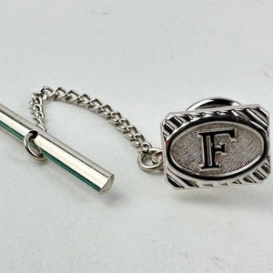 May include: Silver tie tack with a chain and a rectangular design featuring a raised oval with the letter "F" inside.