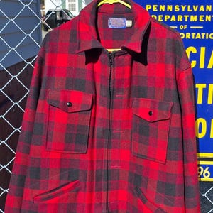 Op de afbeelding: Een rood-zwarte geruite jas met ritssluiting, twee borstzakken en twee zijzakken. De jas hangt aan een gele hanger. De jas heeft een kraag en een label op de bovenrug.