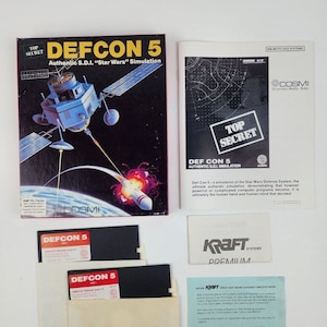 Könnte beinhalten: Eine Schachtel für das Computerspiel Defcon 5, eine Simulation des Star Wars Verteidigungssystems. Die Schachtel zeigt ein Schwarzweißbild eines Satelliten und einer Rakete. Die Schachtel ist mit "Top Secret" und "Authentic S.D.I. "Star Wars" Simulation" beschriftet.