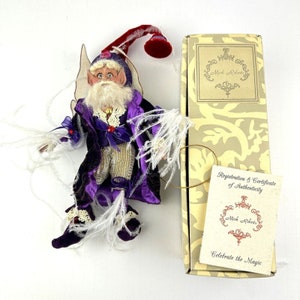 May include: A purple and gold fairy figurine with white feathers, wearing a purple hat and a purple and gold outfit. The figurine is holding a red and gold scepter. The figurine is in a box with a label that says "Mark Roberts Celebrate the Magic".