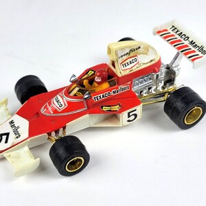 May include: A red, white and gold toy racing car with the words "Texaco-Marlboro" on the side and the number "5" on the front and back. The car has a black spoiler and a driver in the cockpit.