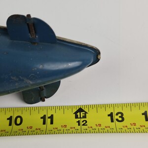 Puede incluir: Un submarino de juguete azul vintage con un ribete plateado y dos peque&ntilde;as aletas. El juguete mide aproximadamente 28 cm de largo, medido por una cinta m&eacute;trica amarilla en la imagen. El juguete muestra signos de desgaste.