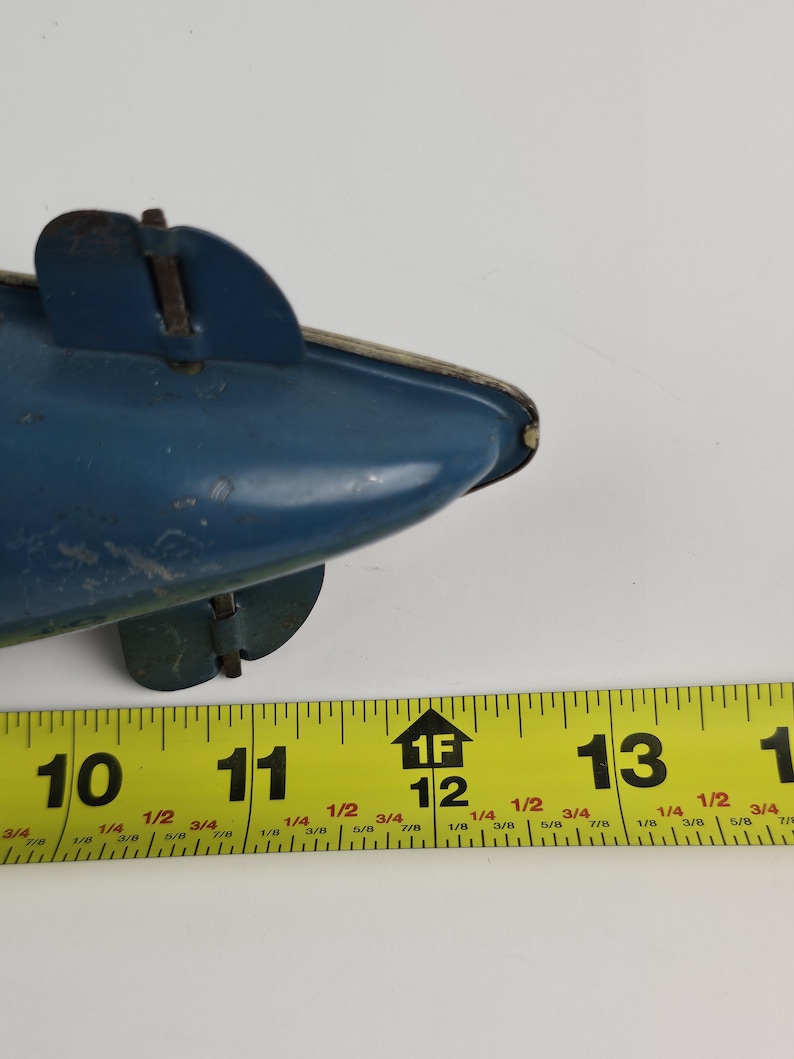 Puede incluir: Un submarino de juguete azul vintage con un ribete plateado y dos peque&ntilde;as aletas. El juguete mide aproximadamente 28 cm de largo, medido por una cinta m&eacute;trica amarilla en la imagen. El juguete muestra signos de desgaste.