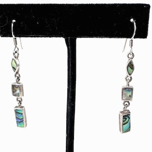 May include: Silver dangle earrings with abalone shell accents. Each earring features a hook, a teardrop-shaped abalone piece, a square abalone piece, and a rectangular abalone piece. The abalone shell has iridescent blue, green, and purple hues.
