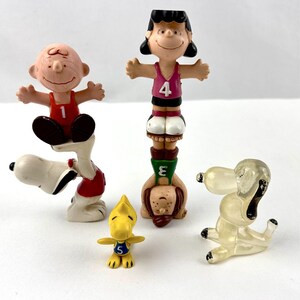 May include: Set of five Peanuts character figurines, including Snoopy, Charlie Brown, Lucy, and Woodstock. The figurines are made of plastic and are painted in various colors. The characters are wearing sports uniforms with numbers on their chests.