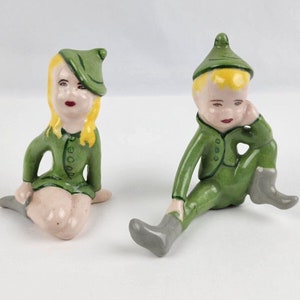 Könnte beinhalten: Zwei Keramikfiguren eines Jungen und eines Mädchens, die grüne Outfits mit passenden Hüten tragen. Der Junge sitzt mit gekreuztem Bein und der Hand am Kinn. Das Mädchen sitzt mit gekreuzten Beinen und den Händen im Schoß.