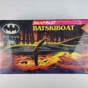 Può includere: Una scatola per un kit di modellismo Bat Skiboat Batman Returns Snapfast. La scatola presenta un'illustrazione in nero e giallo del Bat Skiboat che sfreccia sull'acqua. La scatola presenta anche il logo di Batman Returns e il testo "Snap Together Assembly, No Glue Required".